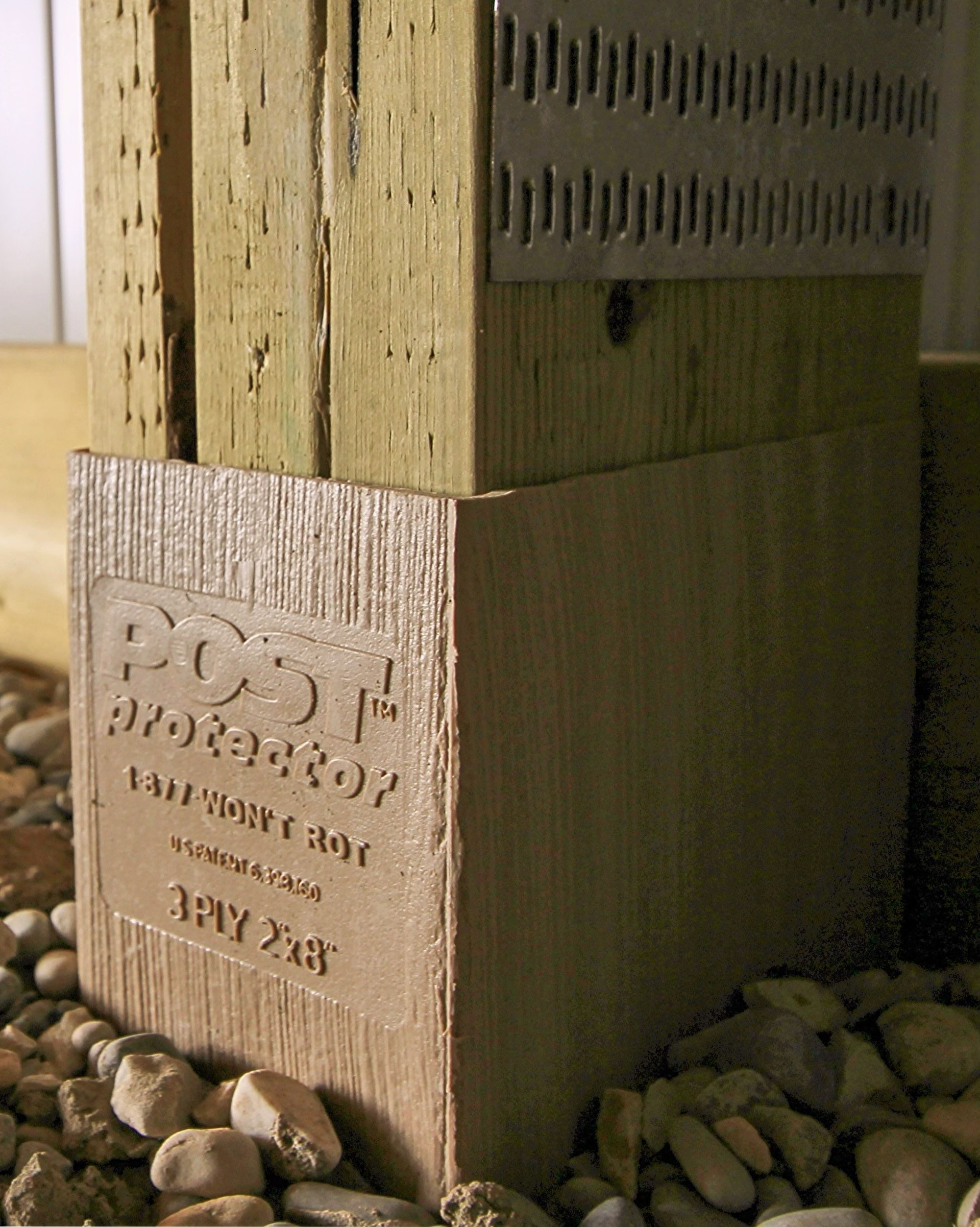 A tan plastic Post Protector sleeve installed at the base of a wooden post surrounded by landscape gravel.
