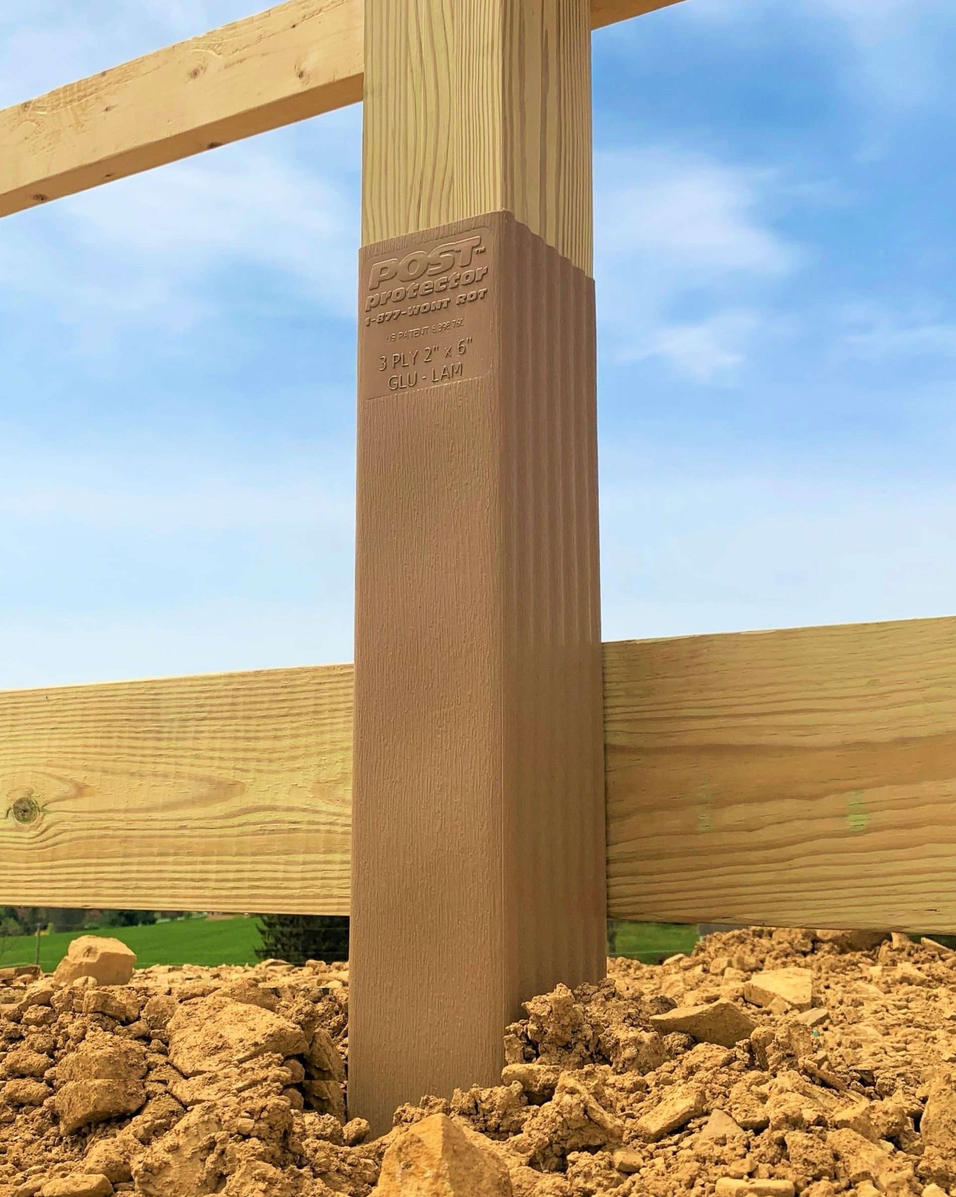 A tan wooden fence post encased in a brown protective sleeve, set in dirt against a blue sky.