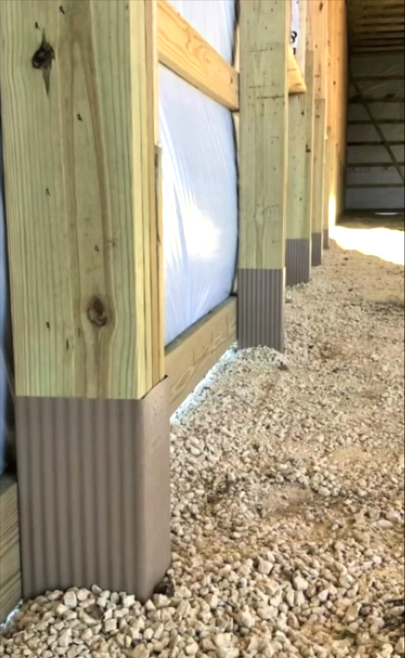 Structural wooden support posts with protective sleeves installed at the base, set against a wall over a gravel ground.