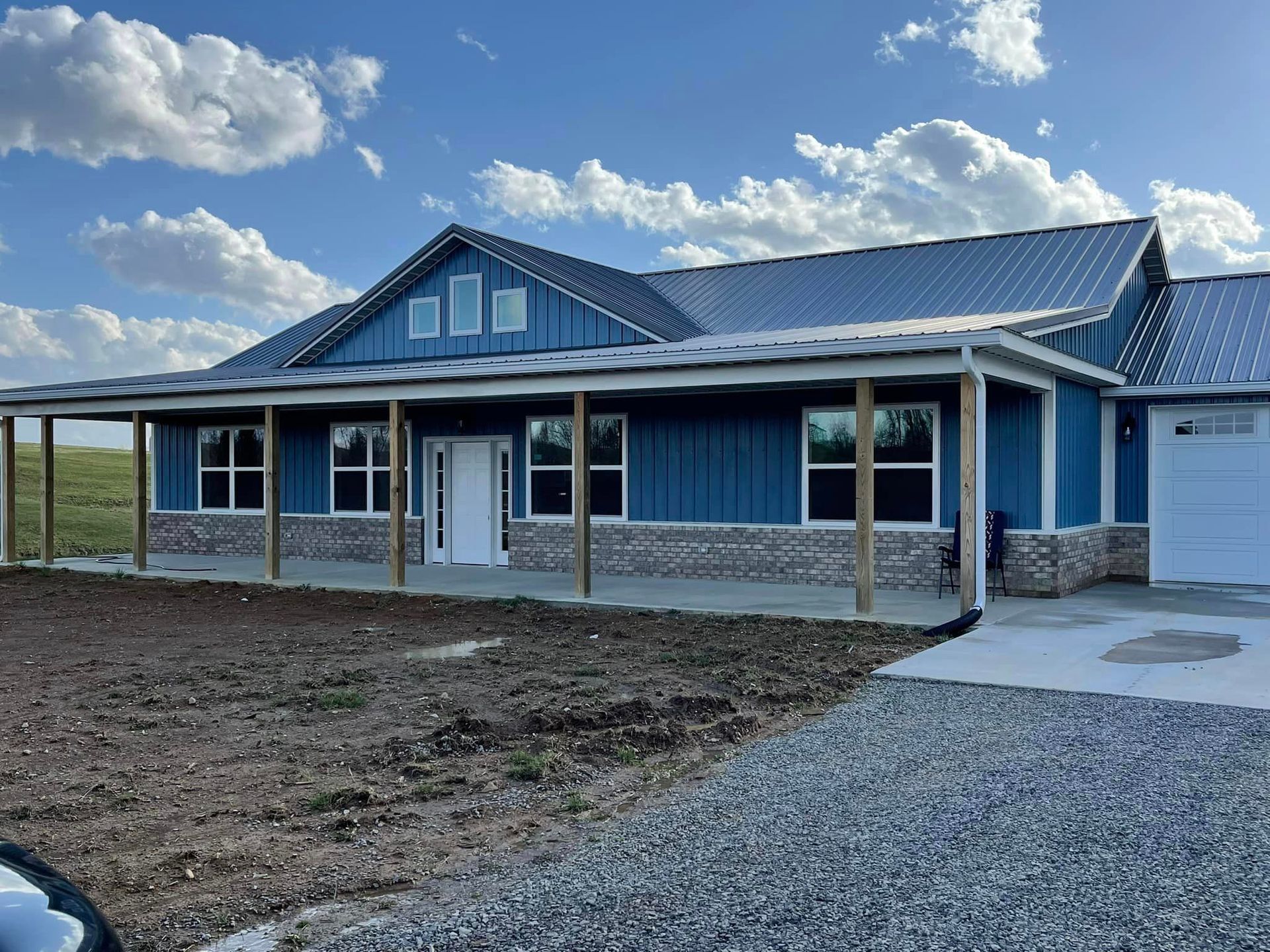 A modern blue metal home with a covered front porch, stone siding, and a gravel driveway under a bright blue sky.