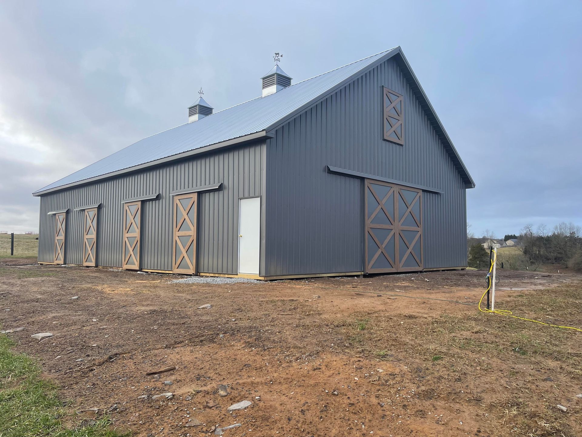 A modern dark grey metal barn with wooden sliding doors and two cupolas, situated on an open, dirt-covered landscape.