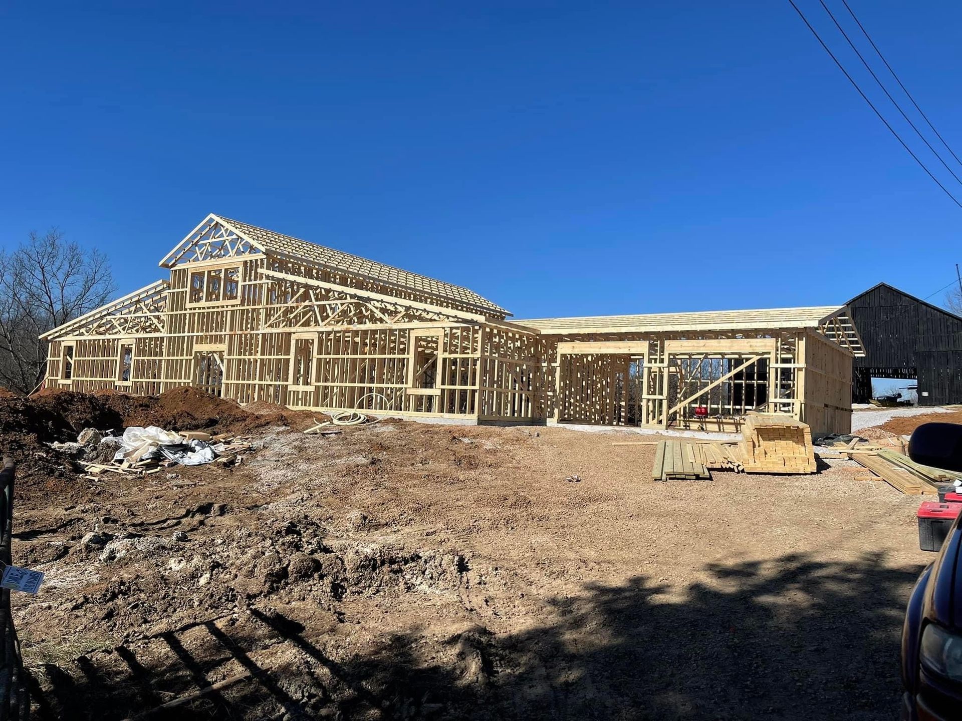 The wooden framework of a house under construction stands on a dirt lot under a clear blue sky.