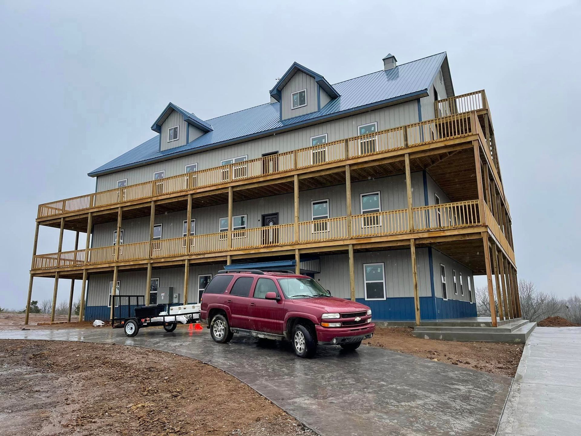 A large gray three-story building with a blue metal roof and wrap-around wooden balconies, with a red SUV parked in front.