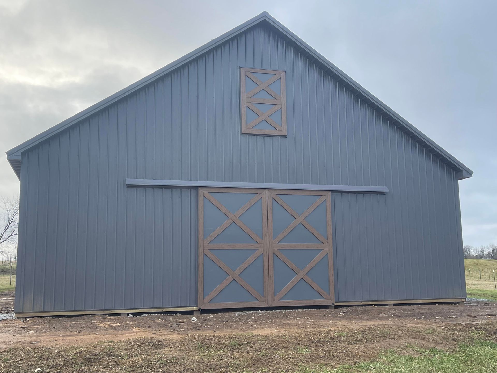 A modern dark blue barn with brown X-patterned double doors and a loft door, set against a cloudy, open field.