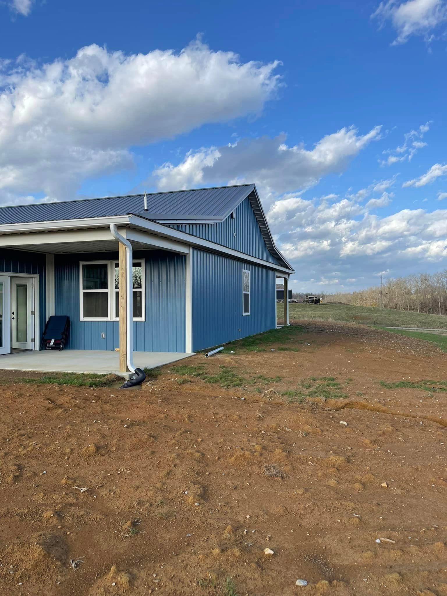 A blue metal-sided house with a covered patio under a partly cloudy sky, set on an undeveloped dirt lot.