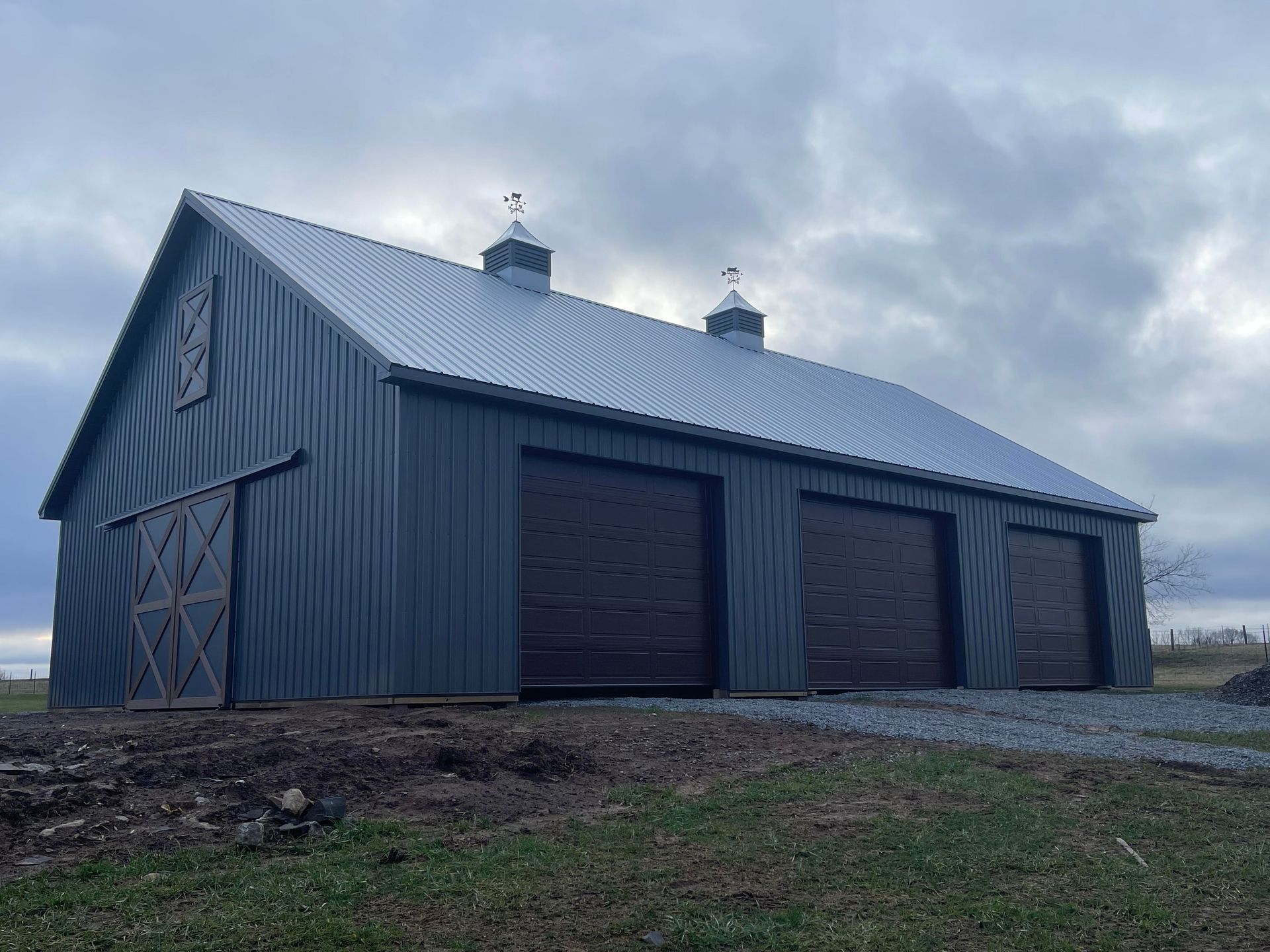 A charcoal-colored metal barn with three garage doors, a sliding side door, and two rooftop cupolas under a cloudy sky.