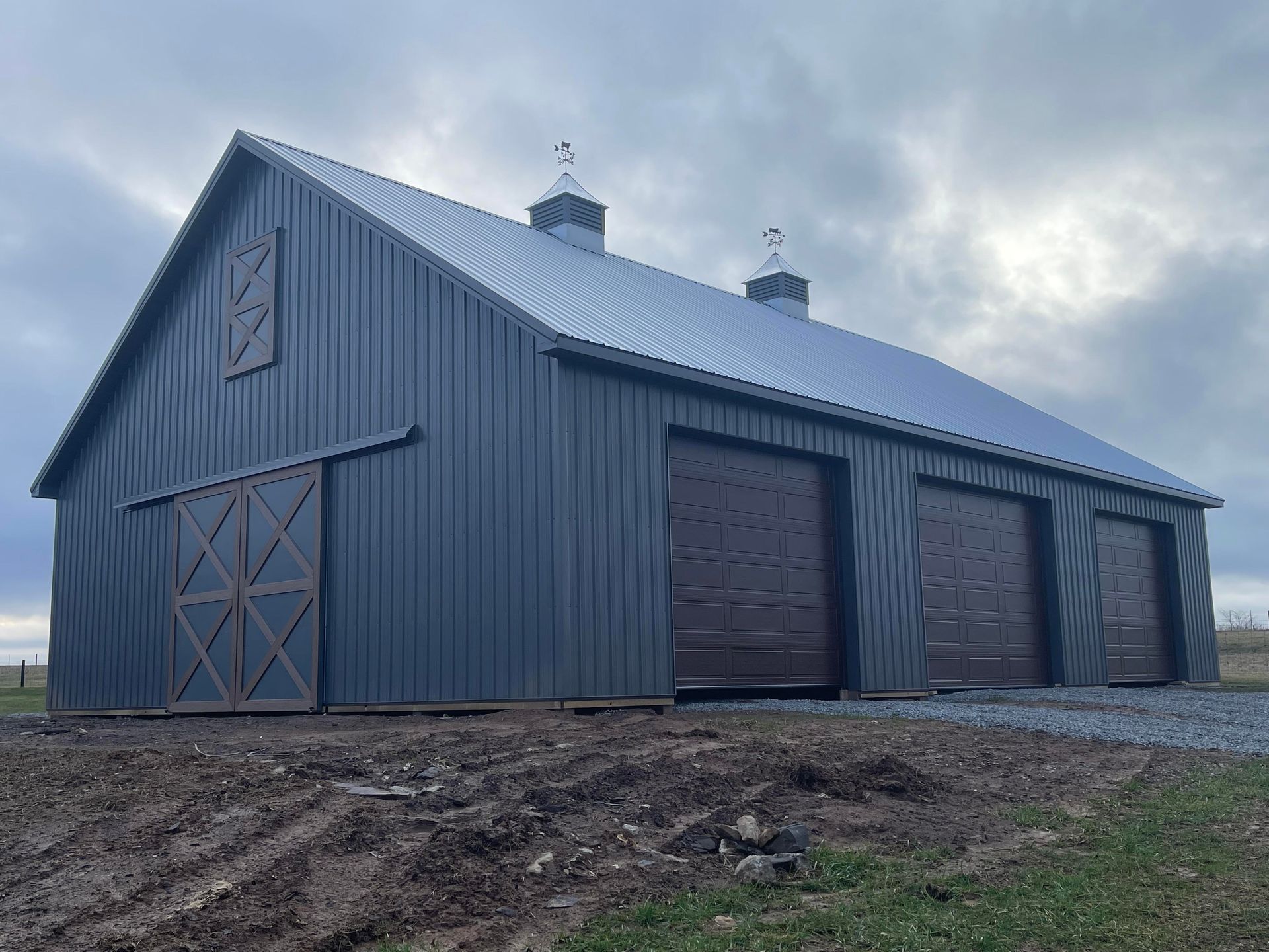 A large, dark gray metal pole barn with a silver roof, sliding barn door, and three garage bays on a gravel lot.