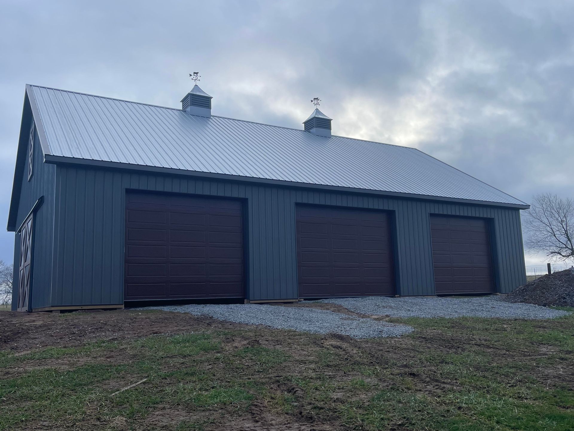 A large, gray metal garage building with three dark brown overhead doors and two cupolas on a gravel lot.