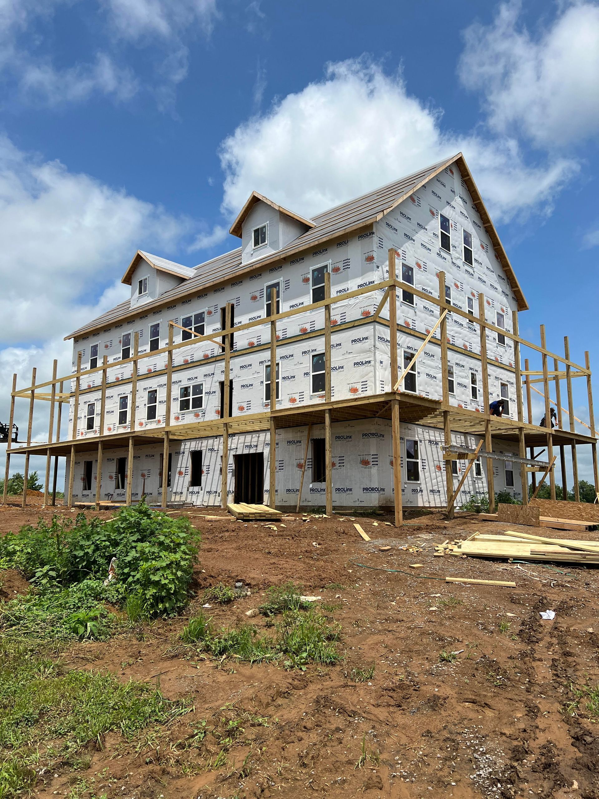 A three-story house under construction, wrapped in white weather-resistant barrier, surrounded by wooden scaffolding.