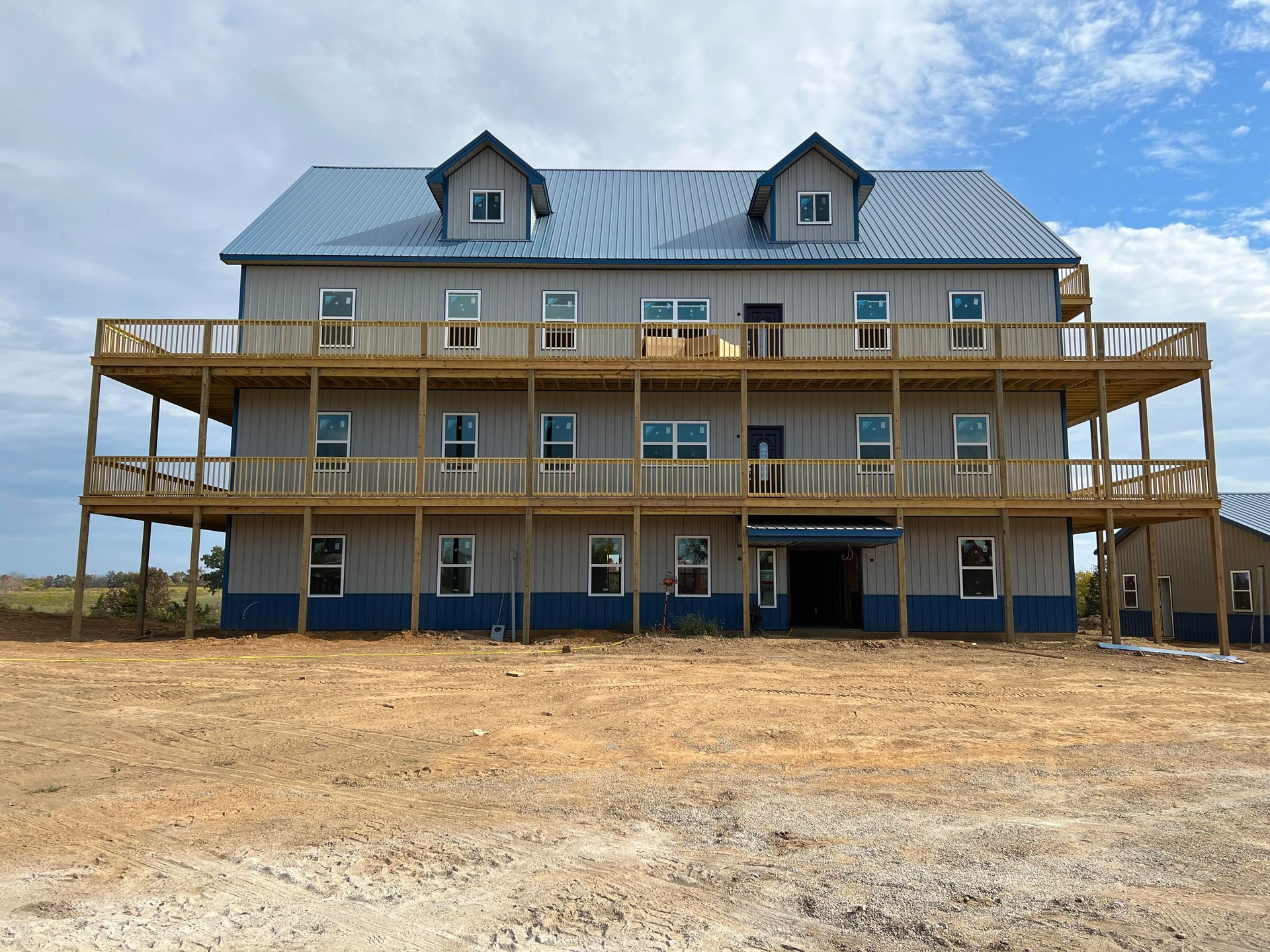 A three-story, grey building with a metal roof and two large wooden wrap-around decks under a cloudy blue sky.