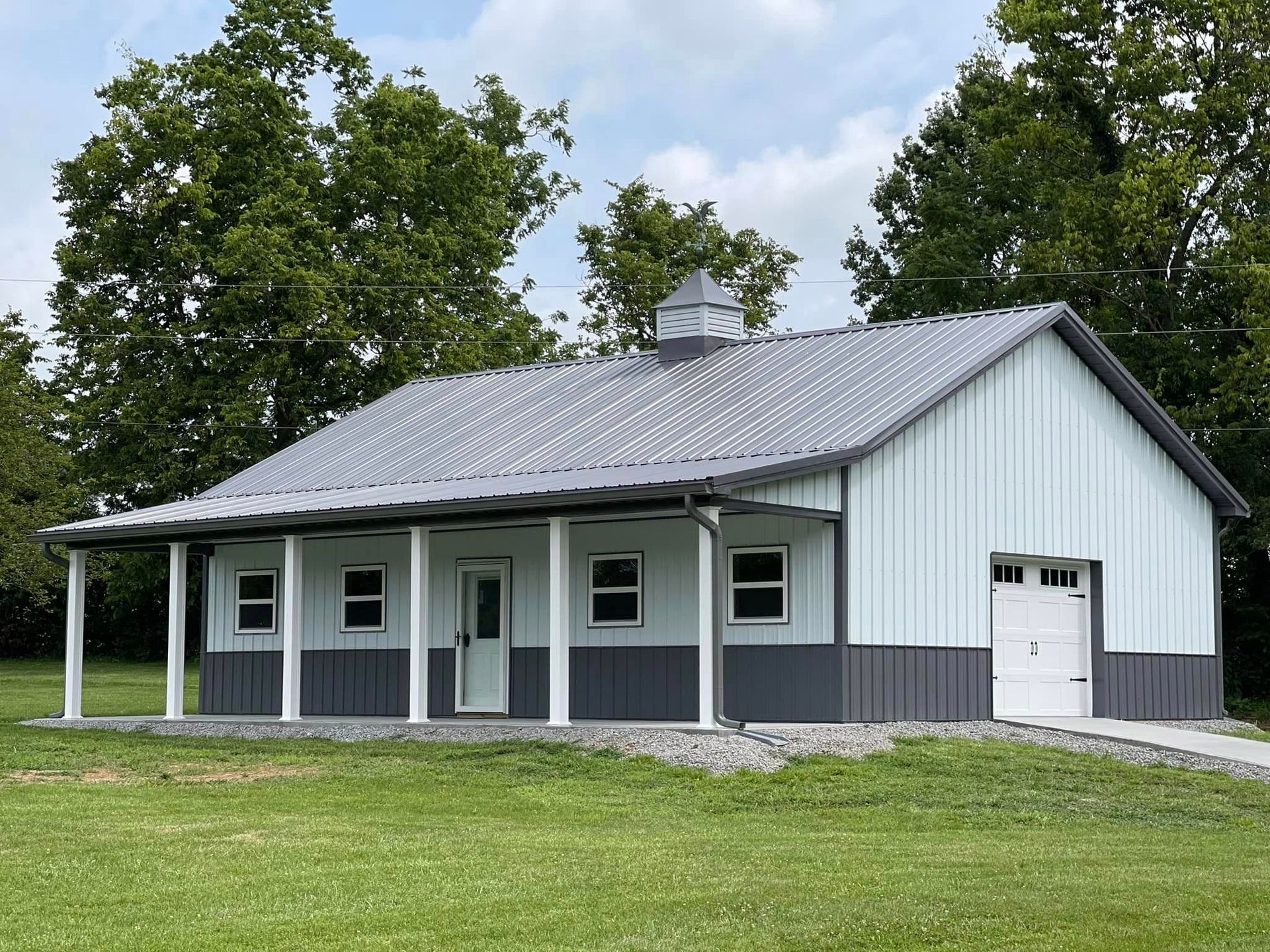 A modern metal barn with a grey roof, light blue siding, a covered porch, and a white garage door in a grassy field.