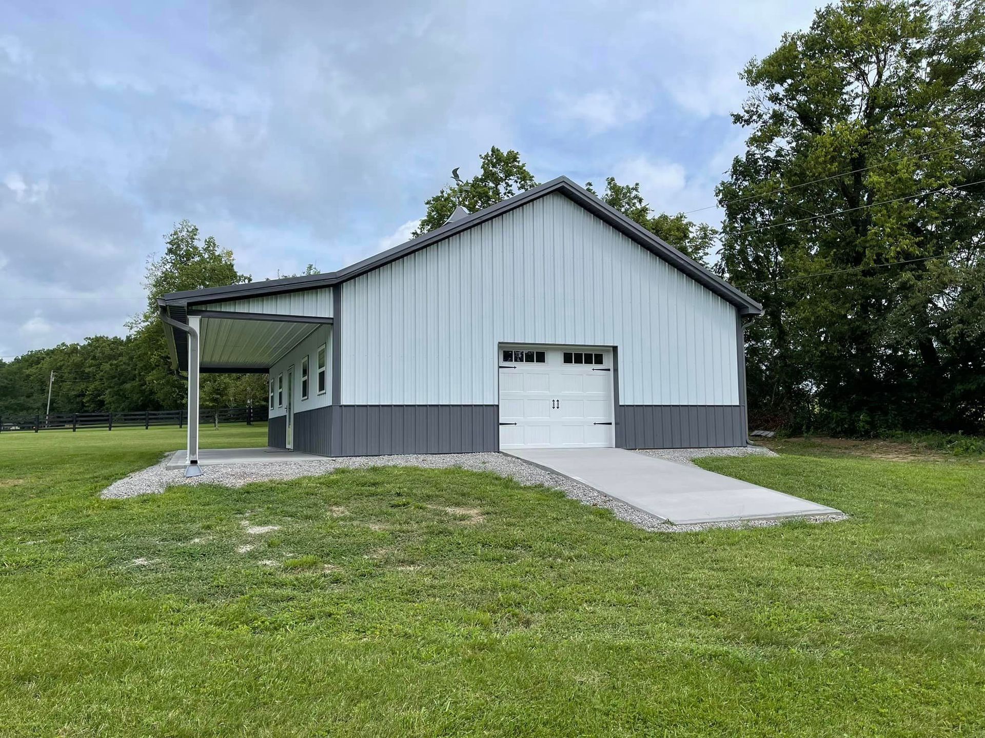 A light gray and dark gray metal garage sits on a grassy field with a white concrete ramp leading to the garage door.