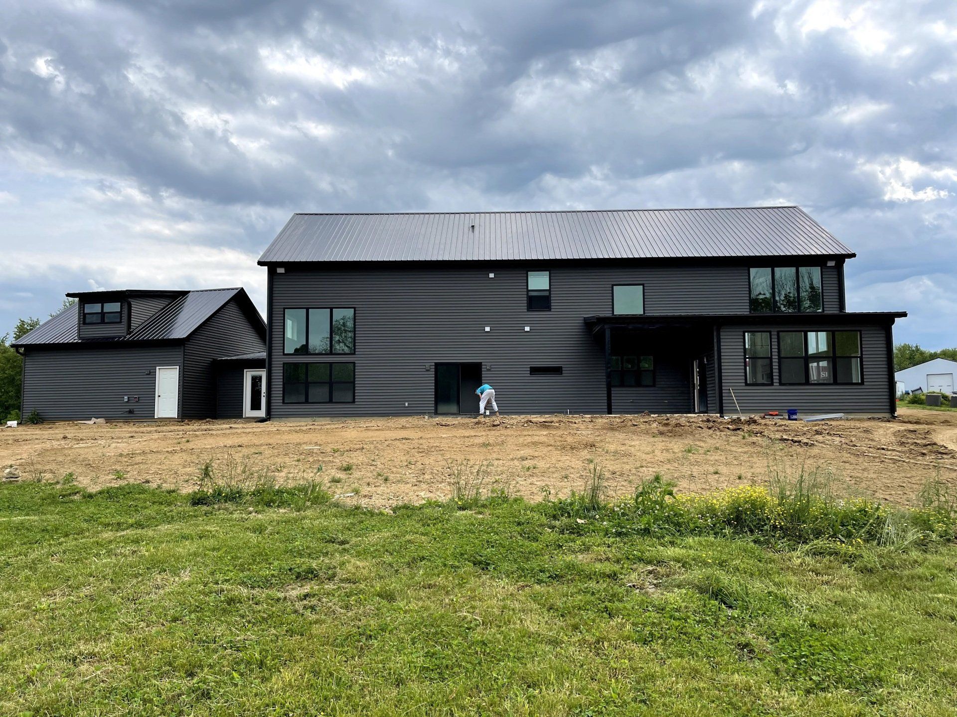 A modern, two-story dark gray house with a metal roof sits on an unfinished dirt lot under a cloudy sky.