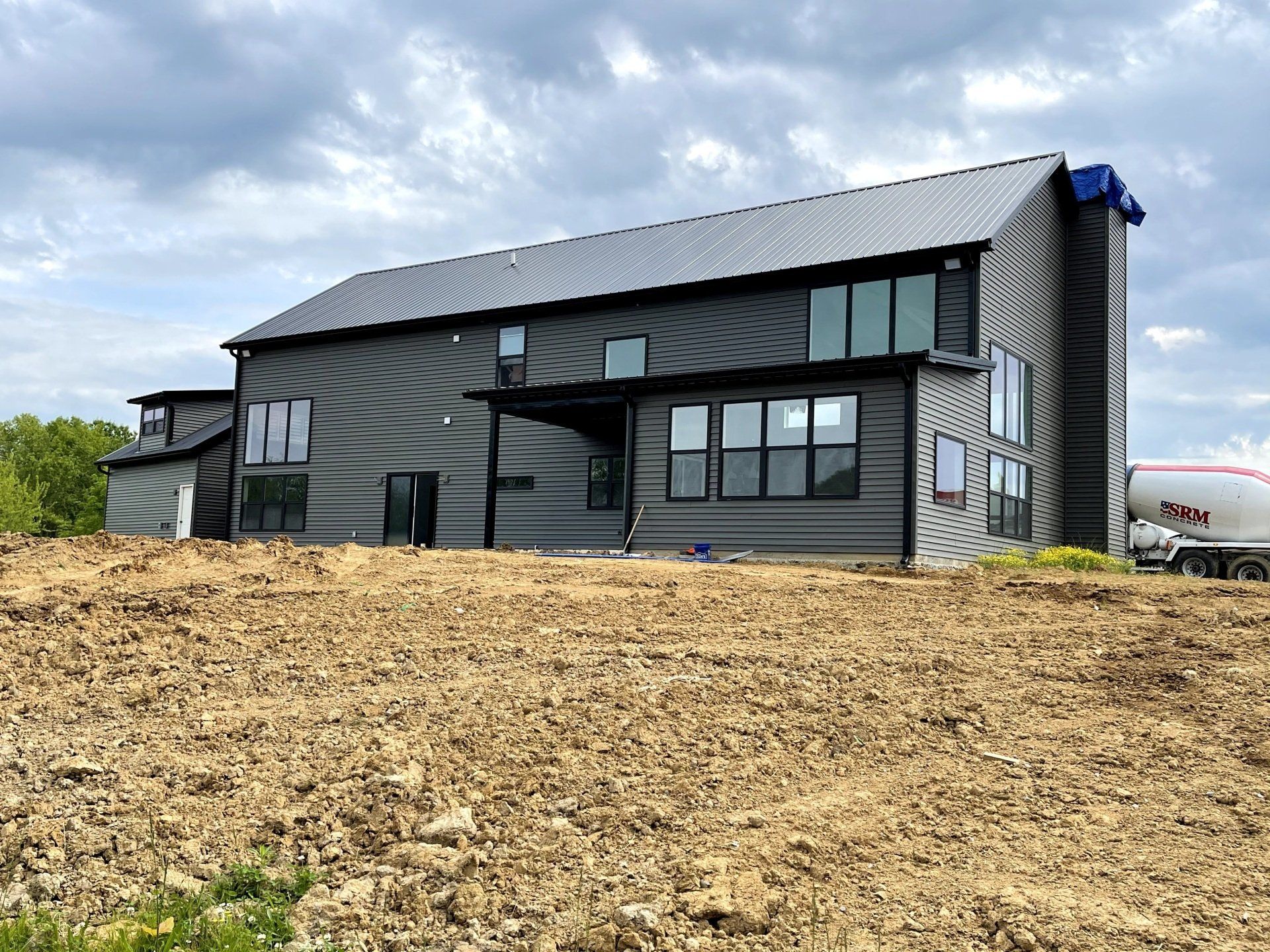 A modern, two-story house with dark gray horizontal siding and a metal roof, under construction on a dirt lot.