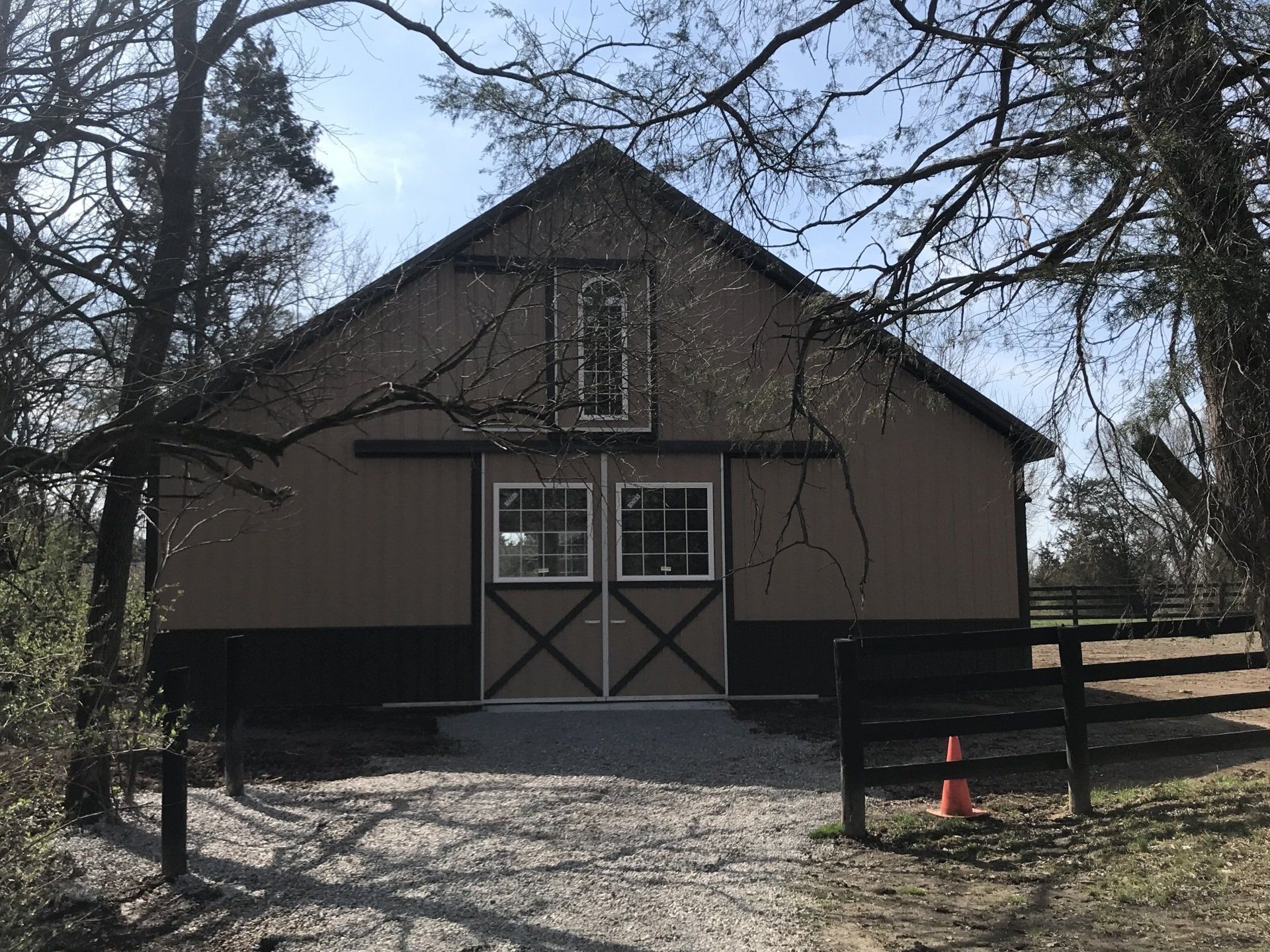 A tan and brown barn with large sliding doors, set in a wooded area with a gravel path and a fence in the foreground.