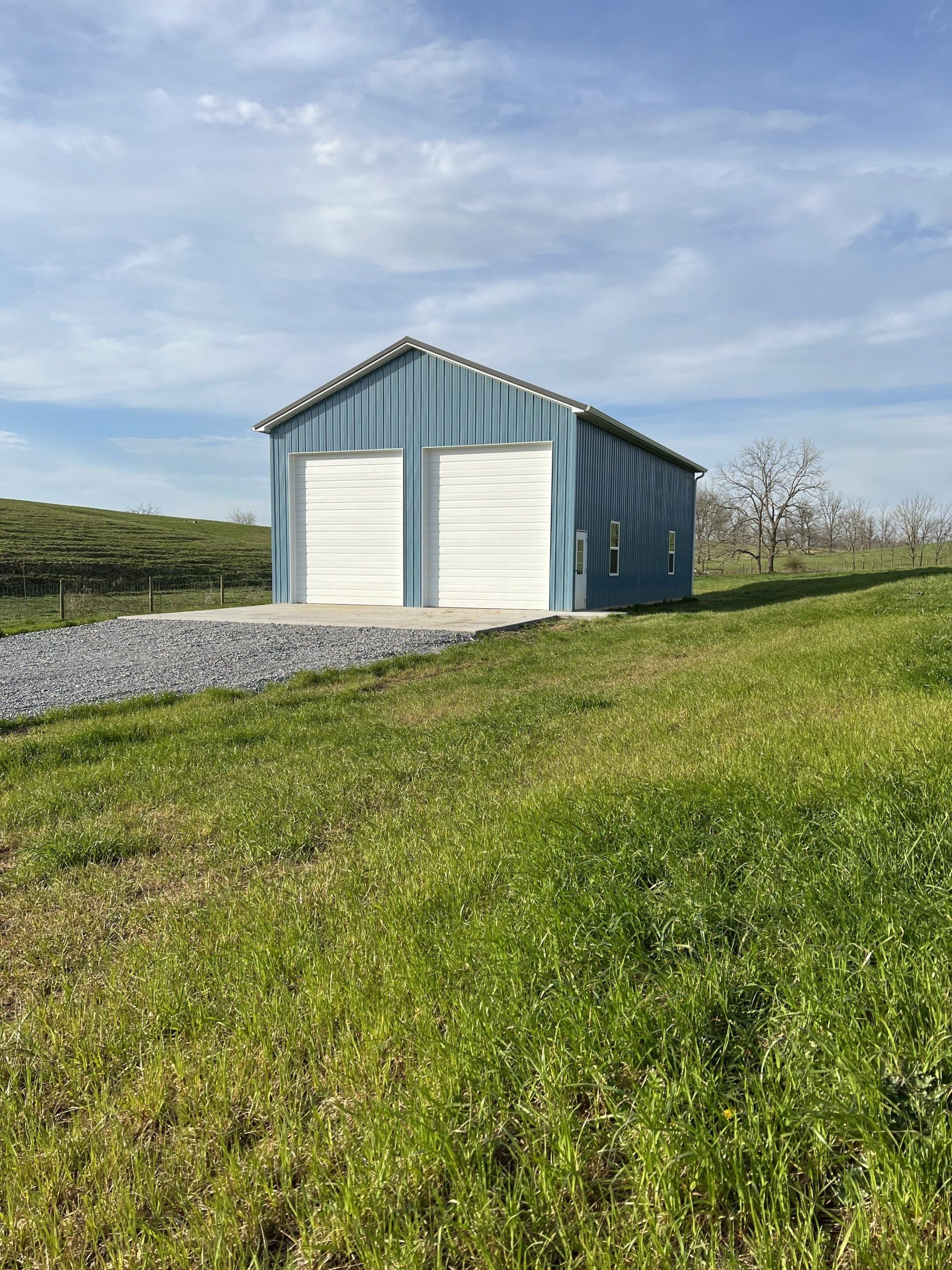 A blue metal garage with two white roll-up doors, set on a gravel pad in a grassy, open field under a sunny sky.