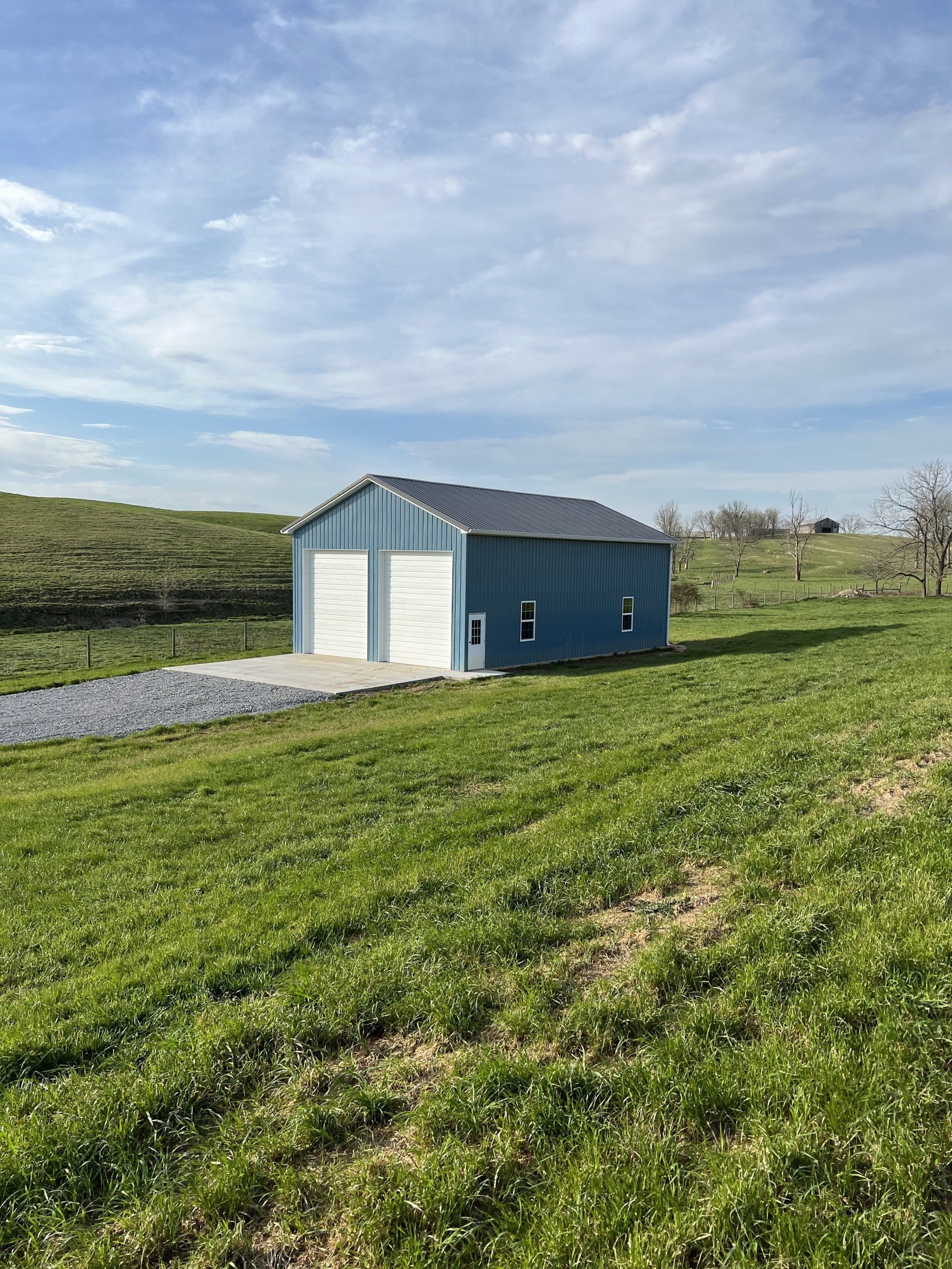 A blue metal garage with two white doors sits on a gravel pad in the middle of a grassy field under a cloudy sky.