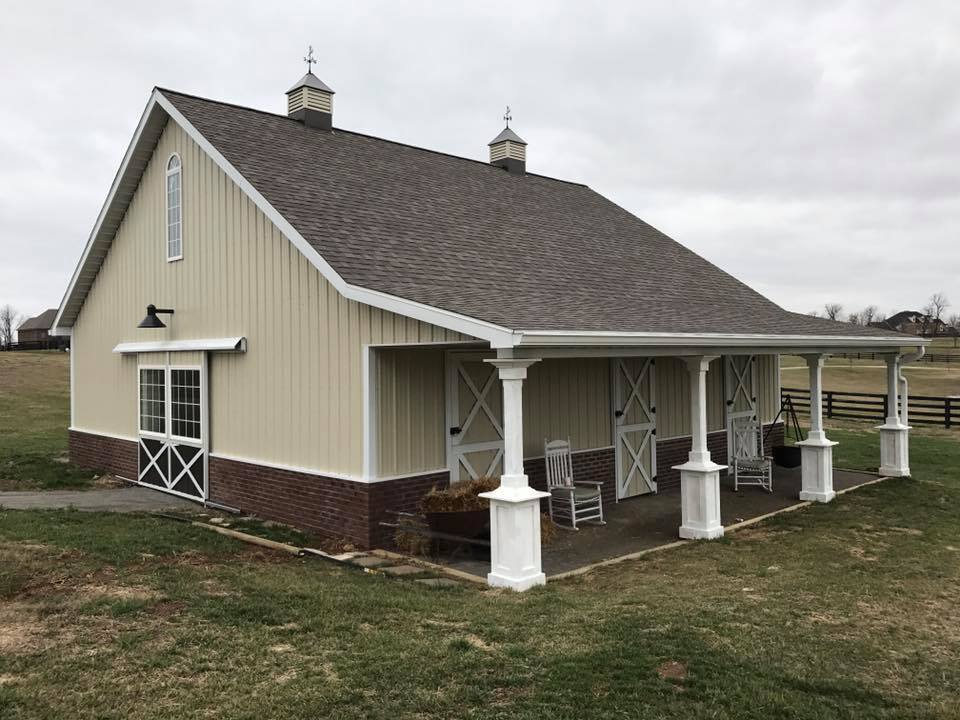 A tan, brick-based barn with a covered porch, white trim, and two cupolas on a grassy landscape under a cloudy sky.