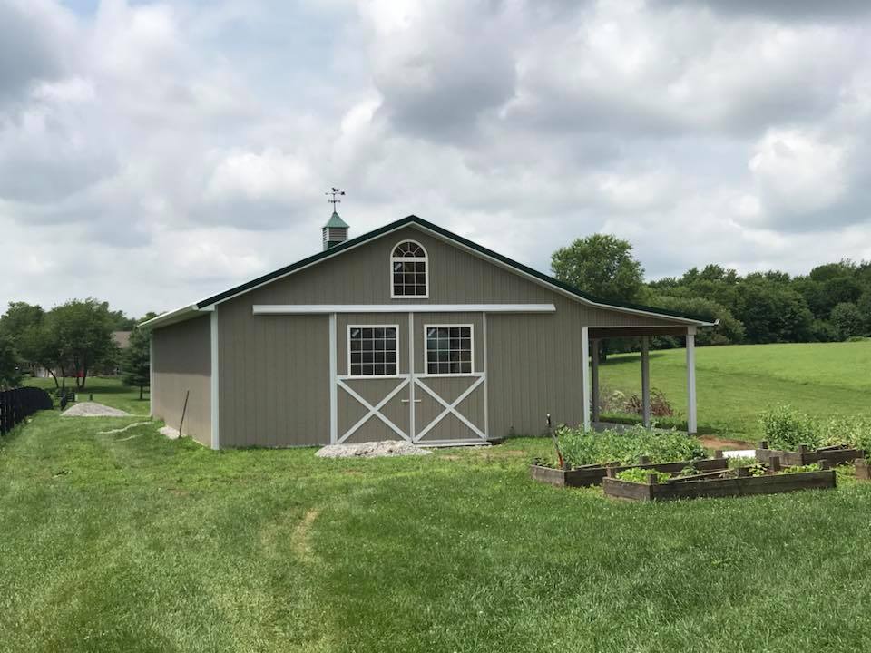A light brown barn with white trim and sliding doors sits in a grassy field under a cloudy sky.
