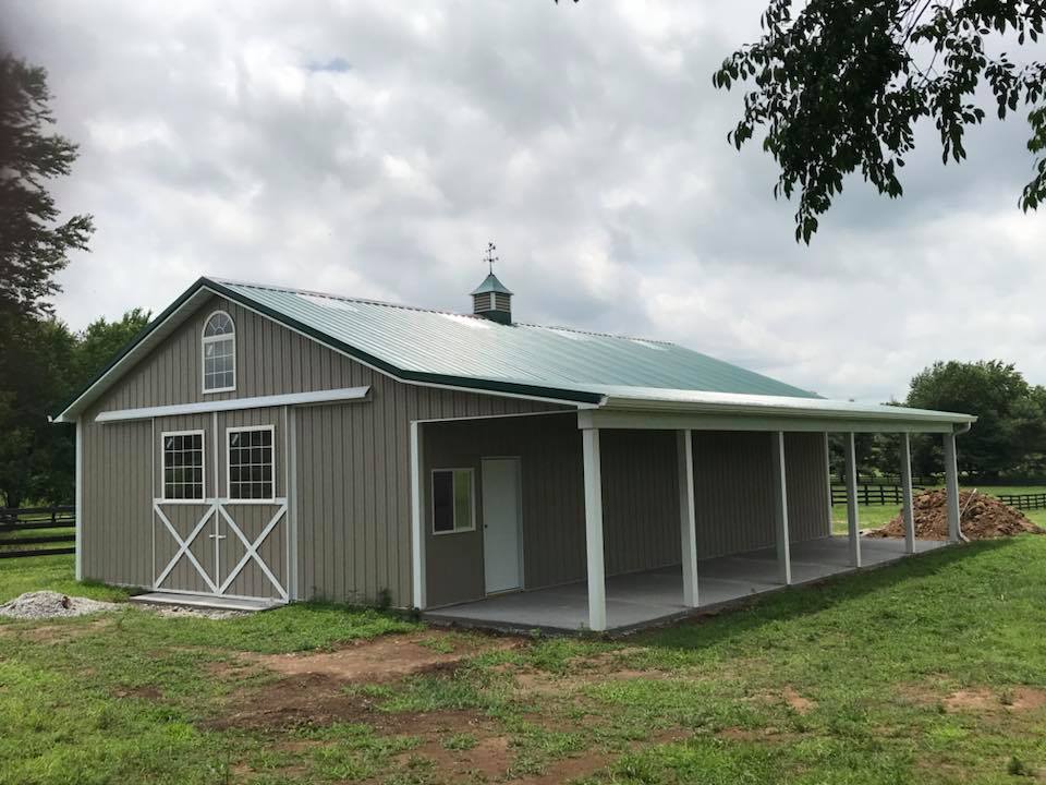 A gray, metal-sided barn with a green roof, a white sliding door, and a covered side porch in a grassy field.