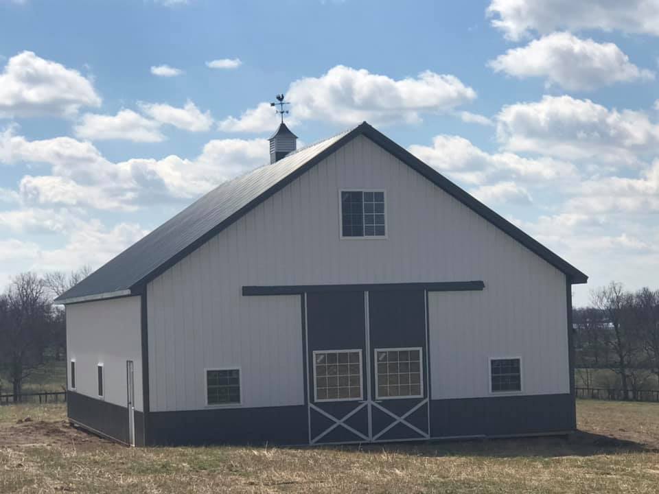 A white and dark gray barn with a black metal roof, a cupola, and sliding doors, set in a grassy field under a blue sky.