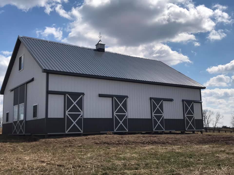 A white and dark gray barn with a metal roof and several sliding doors in a field under a cloudy blue sky.