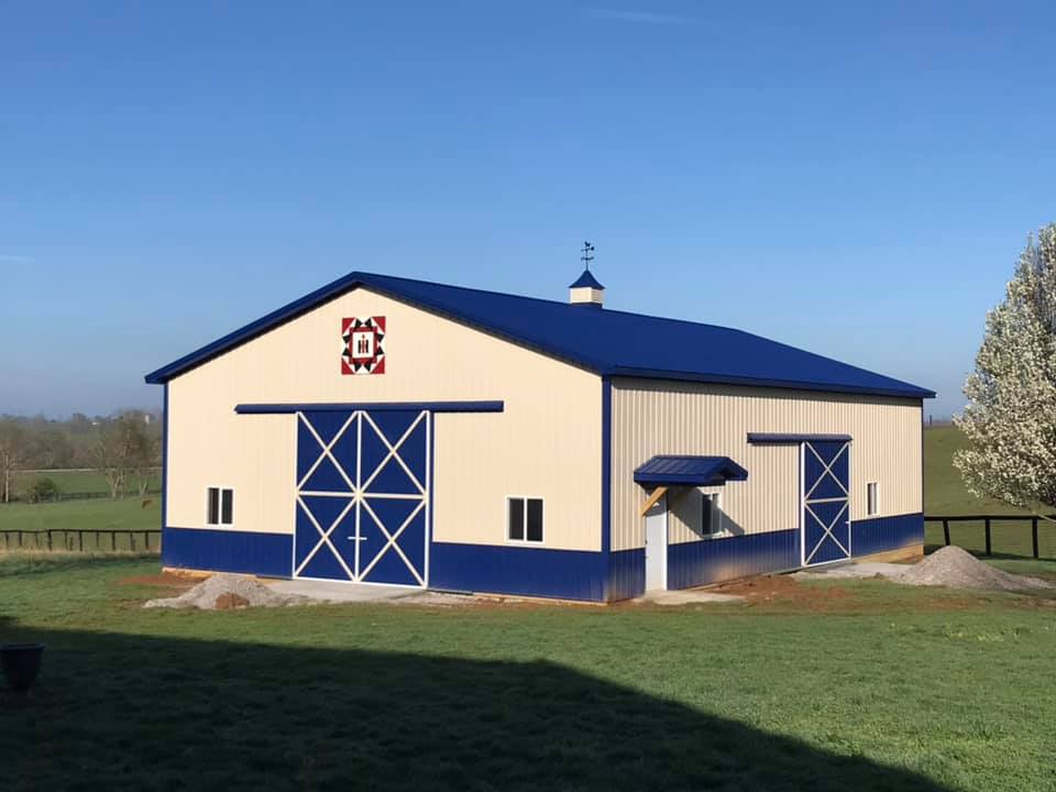 A cream-colored metal barn with a blue roof, trim, and sliding doors, set in a grassy field under a clear blue sky.