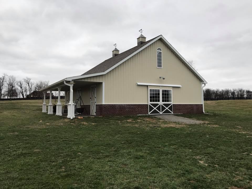 A tan barn with a dark roof and brick base, featuring a porch with white columns, set in a grassy field under a grey sky.