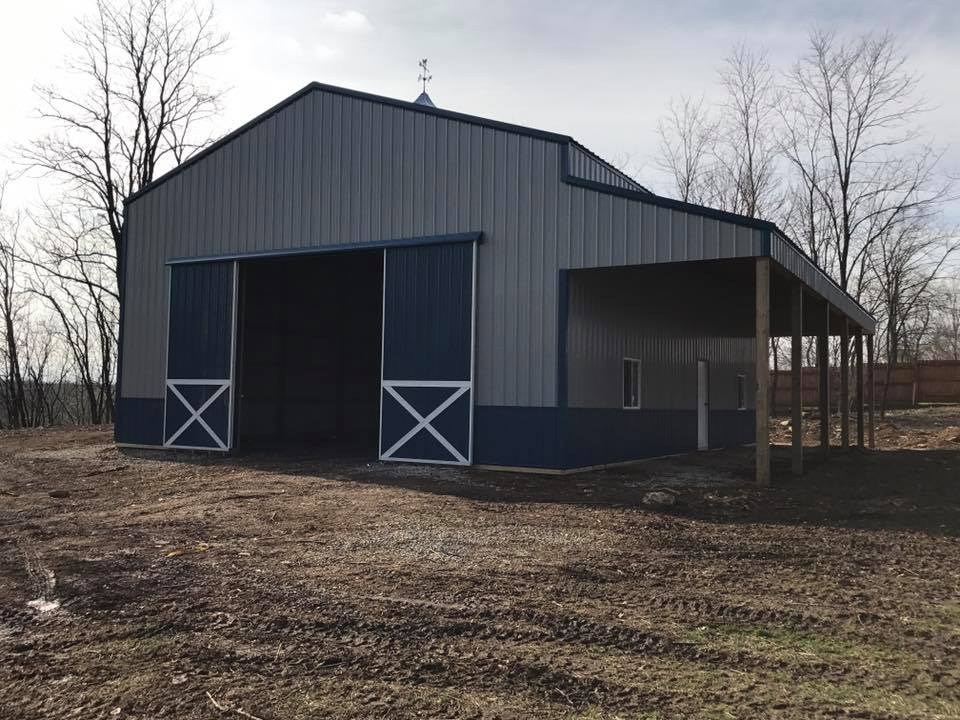A grey and blue metal barn with a sliding front door and an open-sided lean-to, set in a dirt field with bare trees.