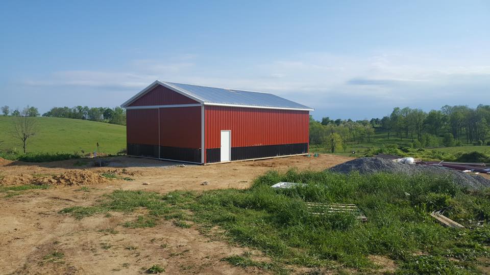 A red pole barn with a metal roof, white trim, and a white door situated in a grassy field under a clear blue sky.