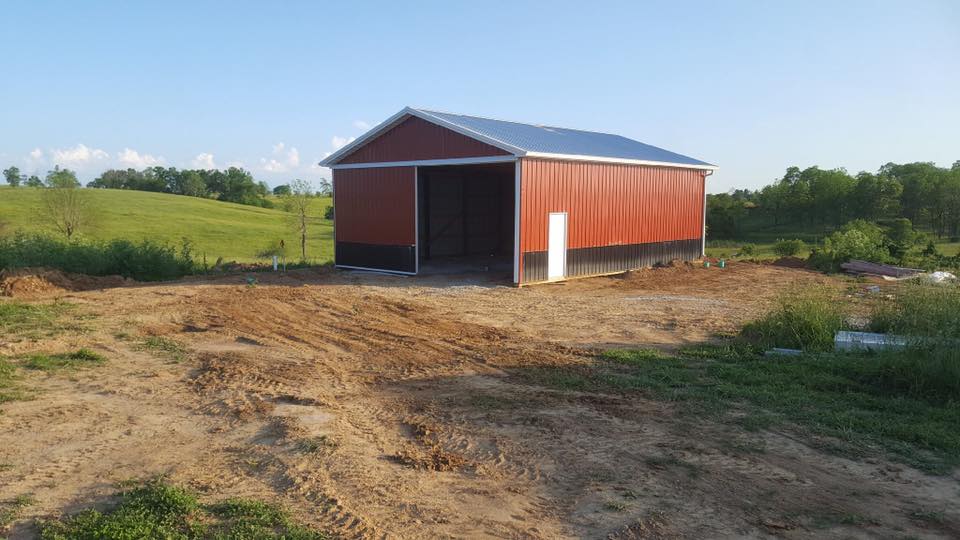 A red barn with a metal roof and open entrance sits on a dirt lot amidst a grassy, rolling landscape under a clear sky.
