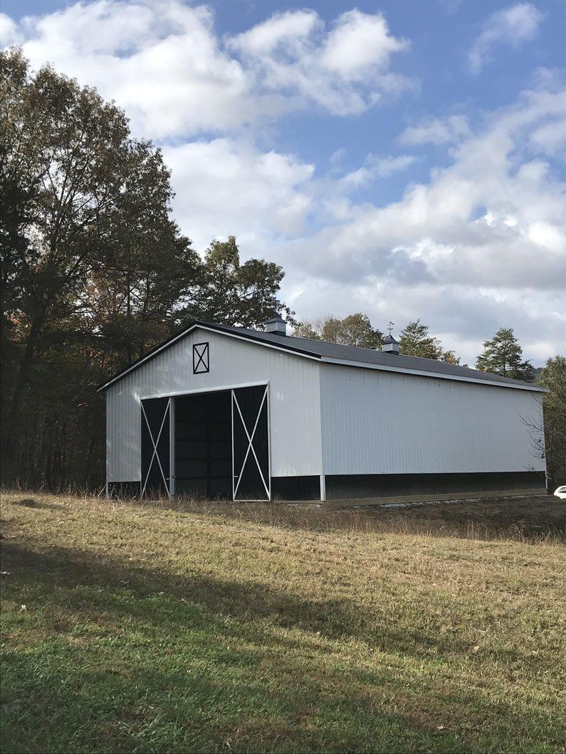 A white pole barn with black trim and cross-braced sliding doors sits in a grassy field surrounded by trees.