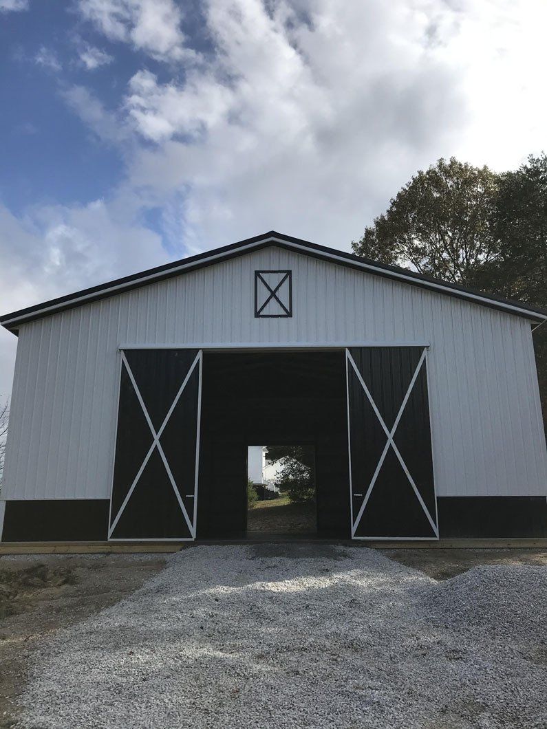 A white barn with black trim and large X-braced sliding doors, situated outdoors on a gravel lot under a cloudy sky.