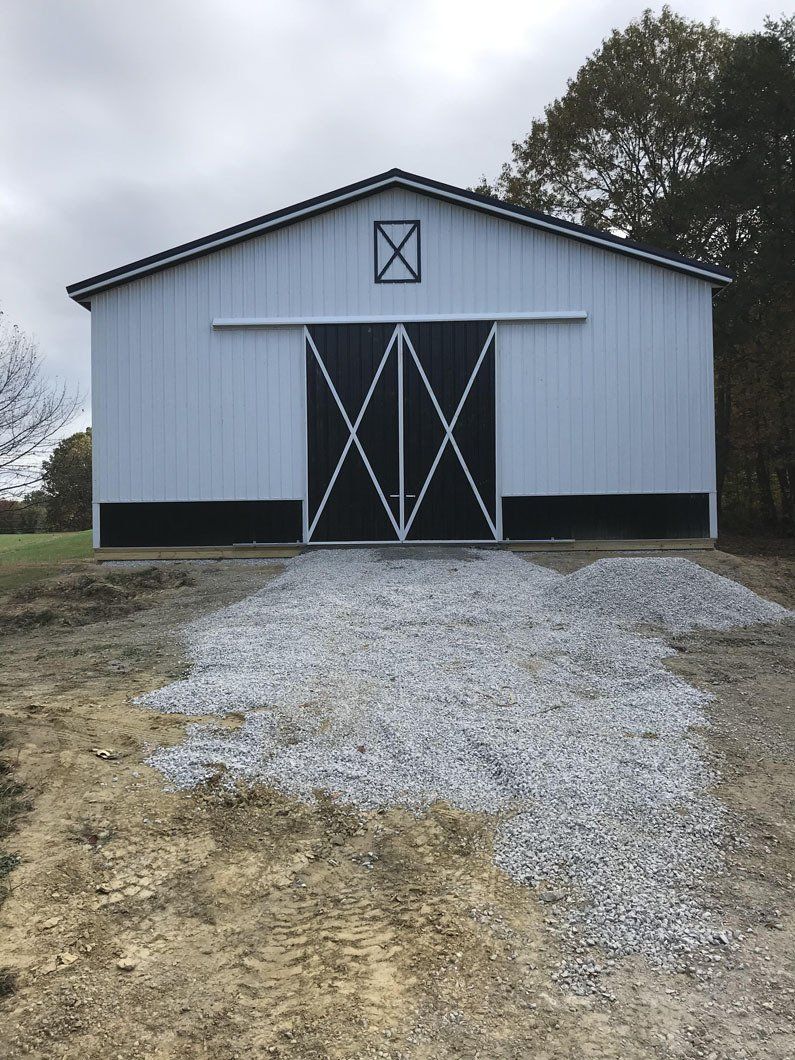 A light-colored pole barn with black sliding doors, an X-pattern design, and a freshly graveled path leading to the entrance.