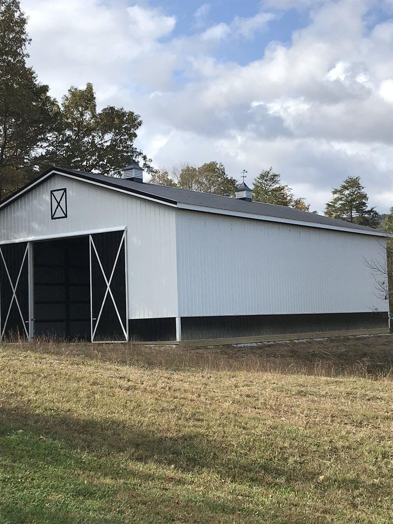 A white, rectangular pole barn with black trim and sliding barn doors stands in a field under a partly cloudy sky.