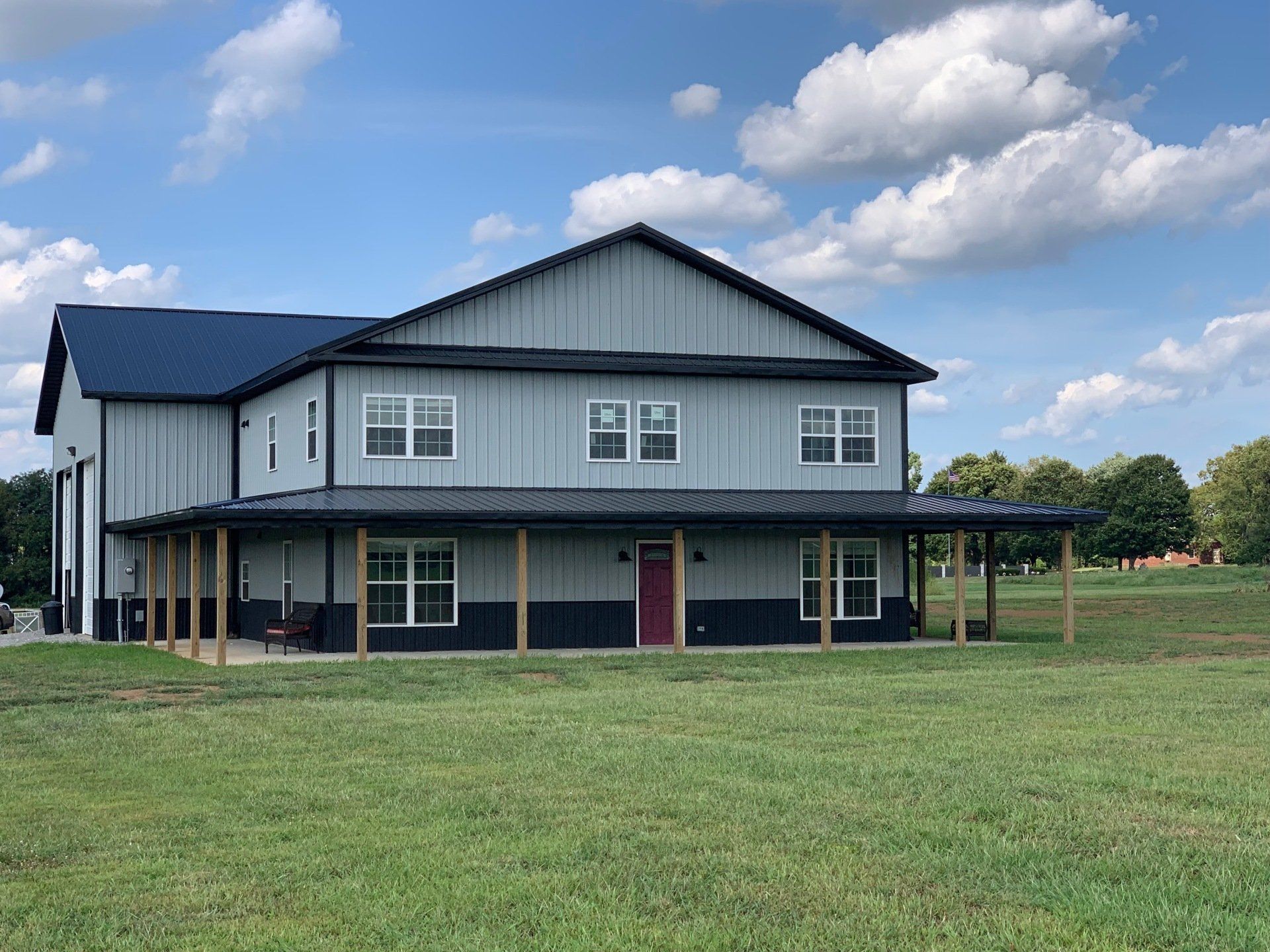 Two-story metal building with gray siding, dark trim, a red door, and a covered porch in a grassy field under a blue sky.