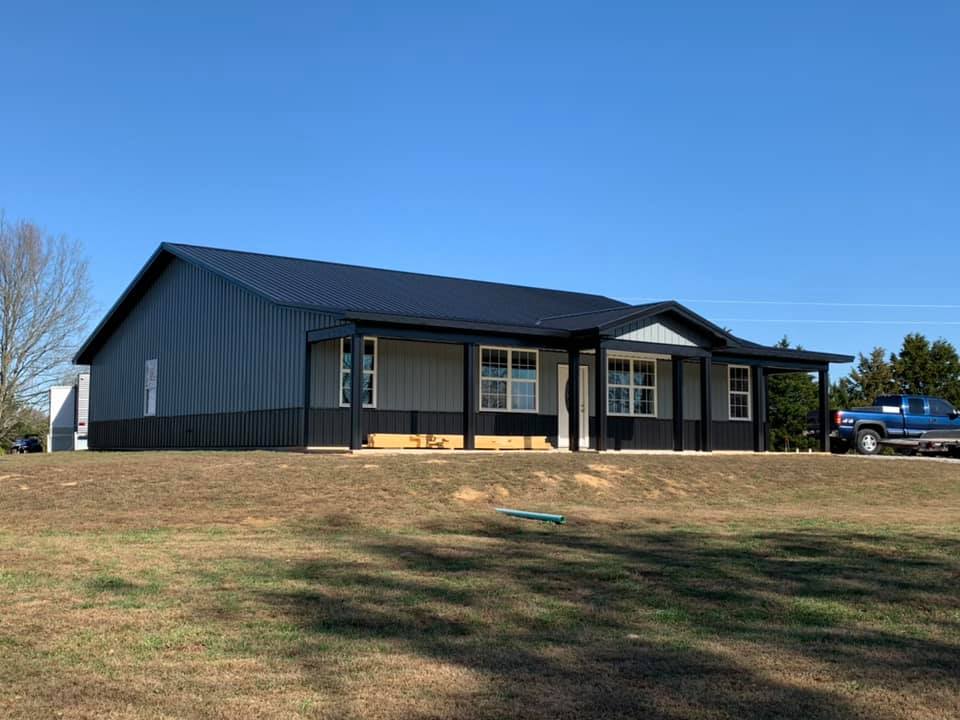 A modern single-story metal building with a dark roof, gray siding, and a covered porch, set on a grassy hill under blue sky.
