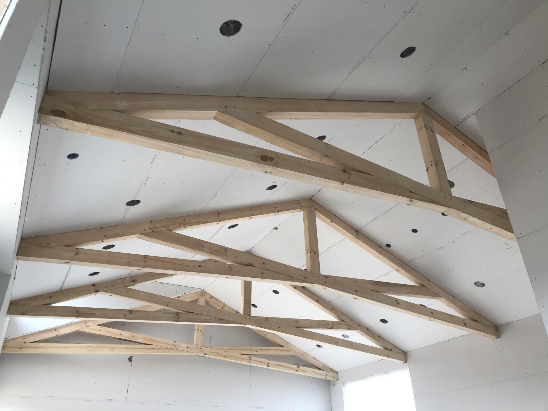 Wooden trusses supporting a vaulted ceiling with installed drywall and recessed light cutouts.