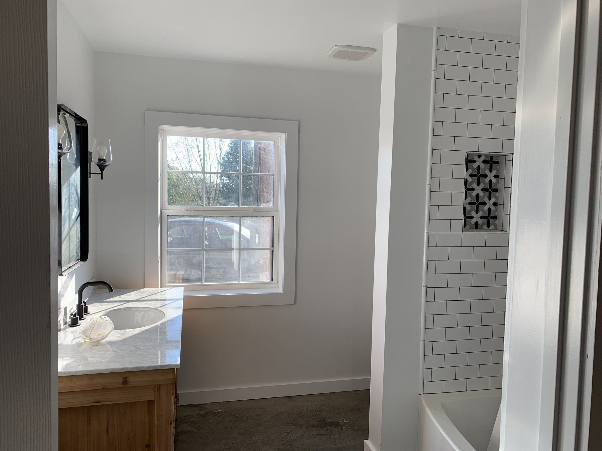 A bathroom with a wood vanity, white countertop, window, and a tiled shower wall featuring a decorative accent niche.