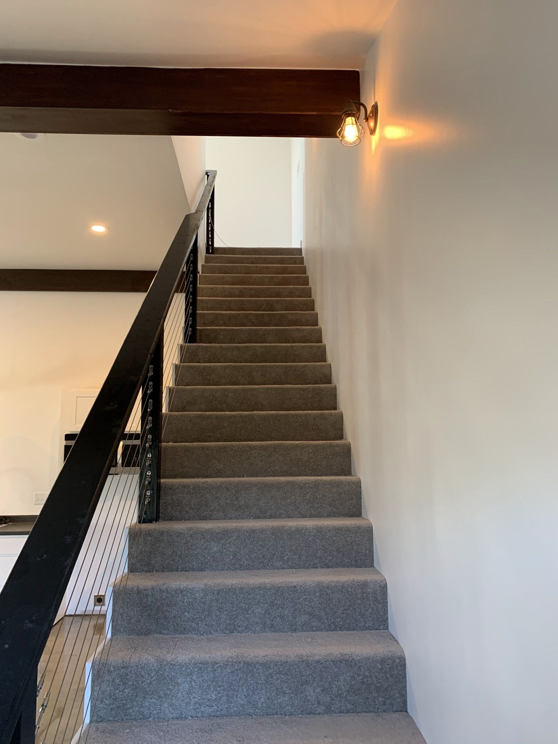 Stairs covered in gray carpet, leading upward toward a landing with a wooden beam above and a wall-mounted light.