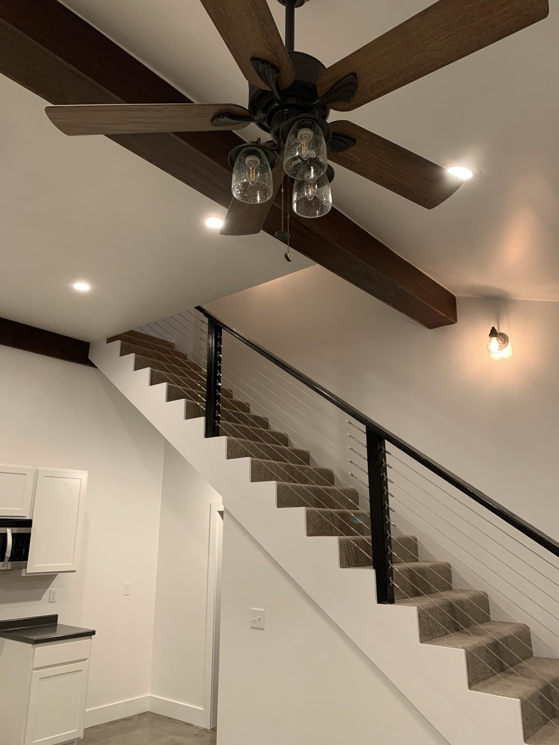 A staircase with black metal railings and cable infill leads upward beneath dark wooden ceiling beams and a ceiling fan.