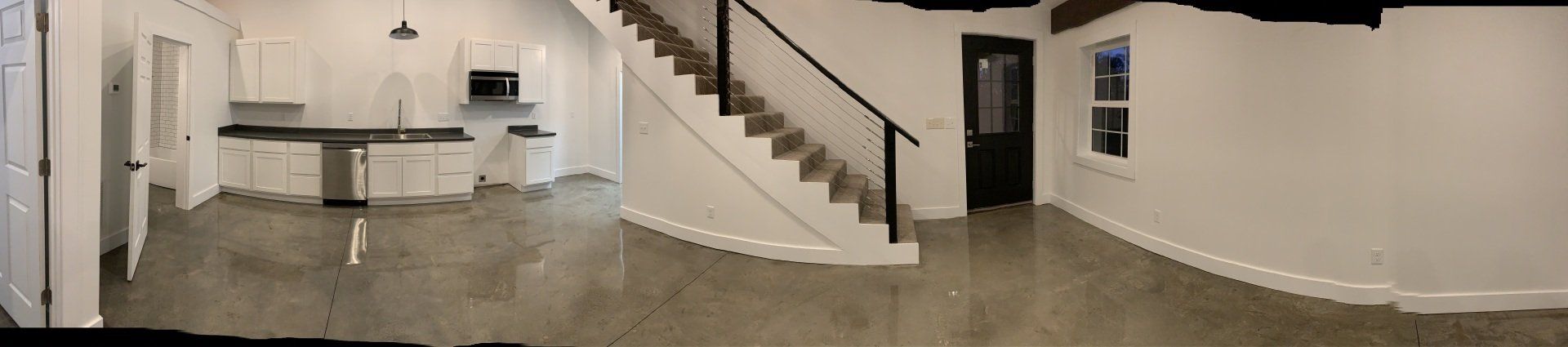 A wide-angle interior view of a modern open-concept space featuring a white kitchen, carpeted stairs, and concrete floors.