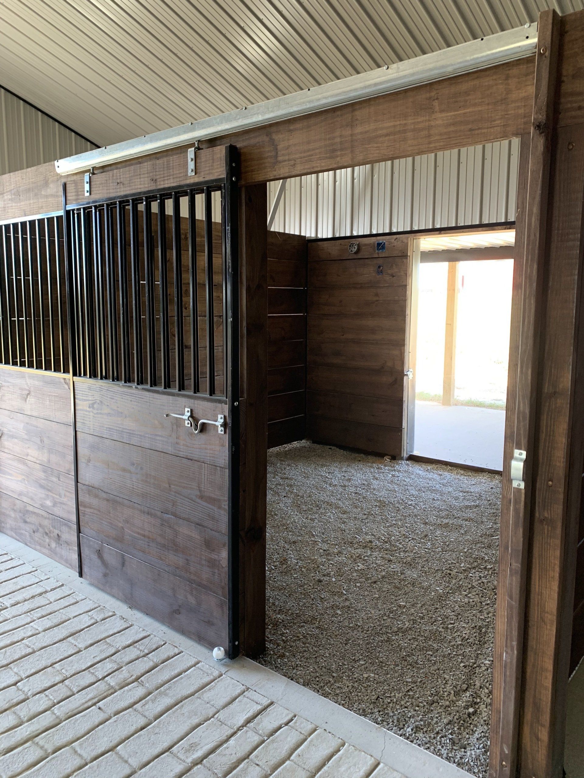 A horse stall with a dark wood sliding door, black metal bars, and a gravel-filled interior in a stable.