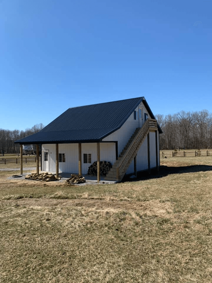 A white barn with a black metal roof, a front porch, and an exterior wooden staircase, set in a grassy rural field.