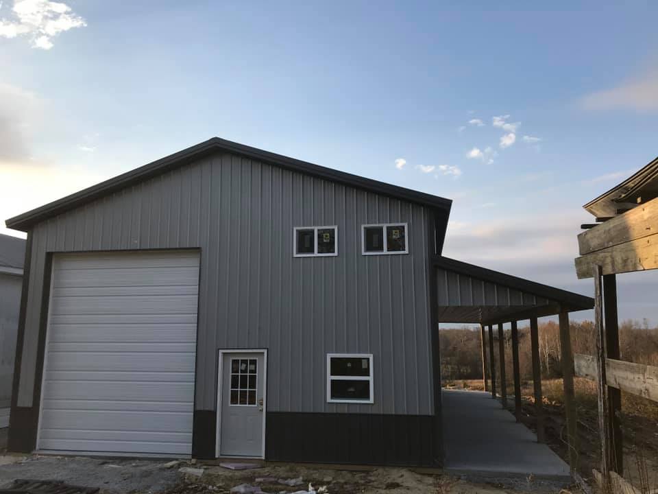 A two-story grey metal building with a white garage door, a side entry door, small windows, and an attached open porch.