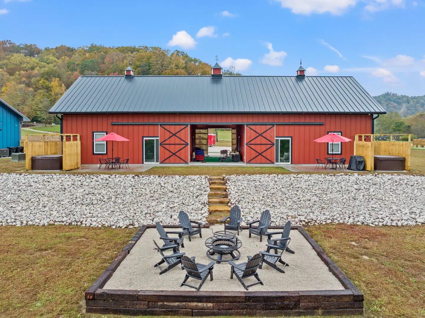 A red barn with a metal roof, outdoor hot tubs, and a gravel fire pit area with chairs in a grassy field.