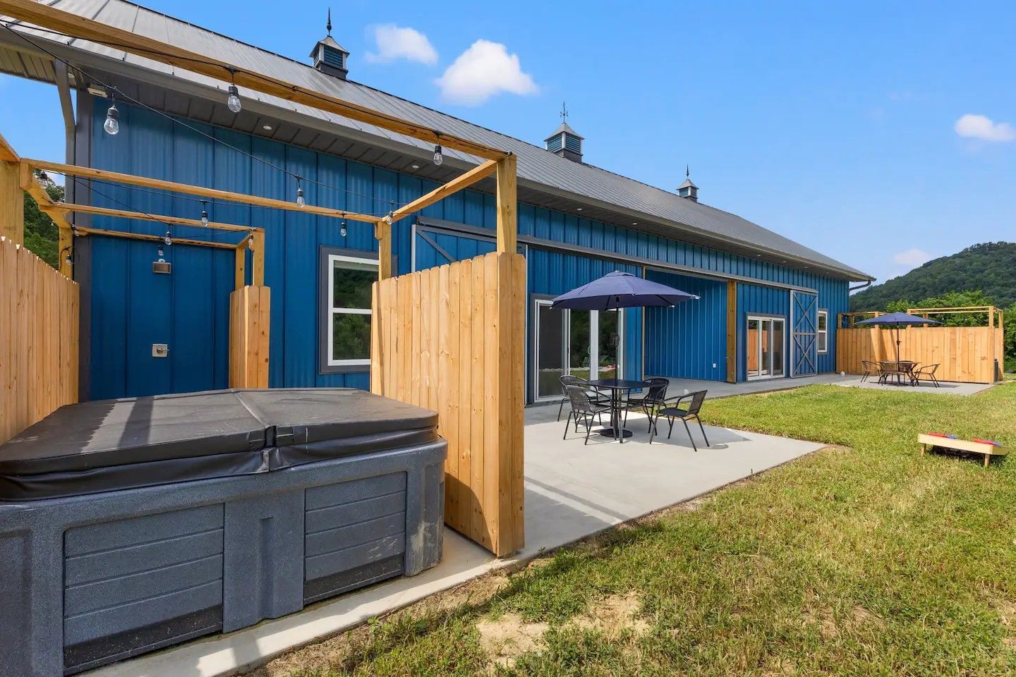A blue metal-sided building with outdoor patios, wooden privacy screens, hot tubs, umbrellas, and dining sets on grass.