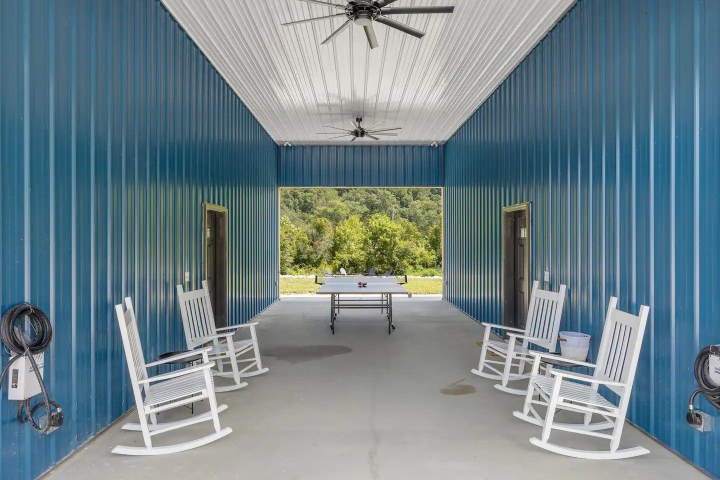 Blue metal-walled covered porch with two rows of white rocking chairs facing a ping pong table, open to a grassy view.
