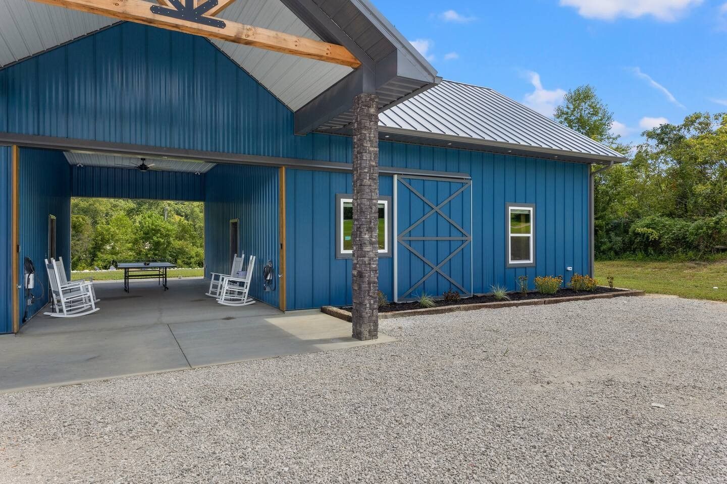 A blue barn-style building with a covered patio, featuring rocking chairs on a concrete floor and a gravel driveway.