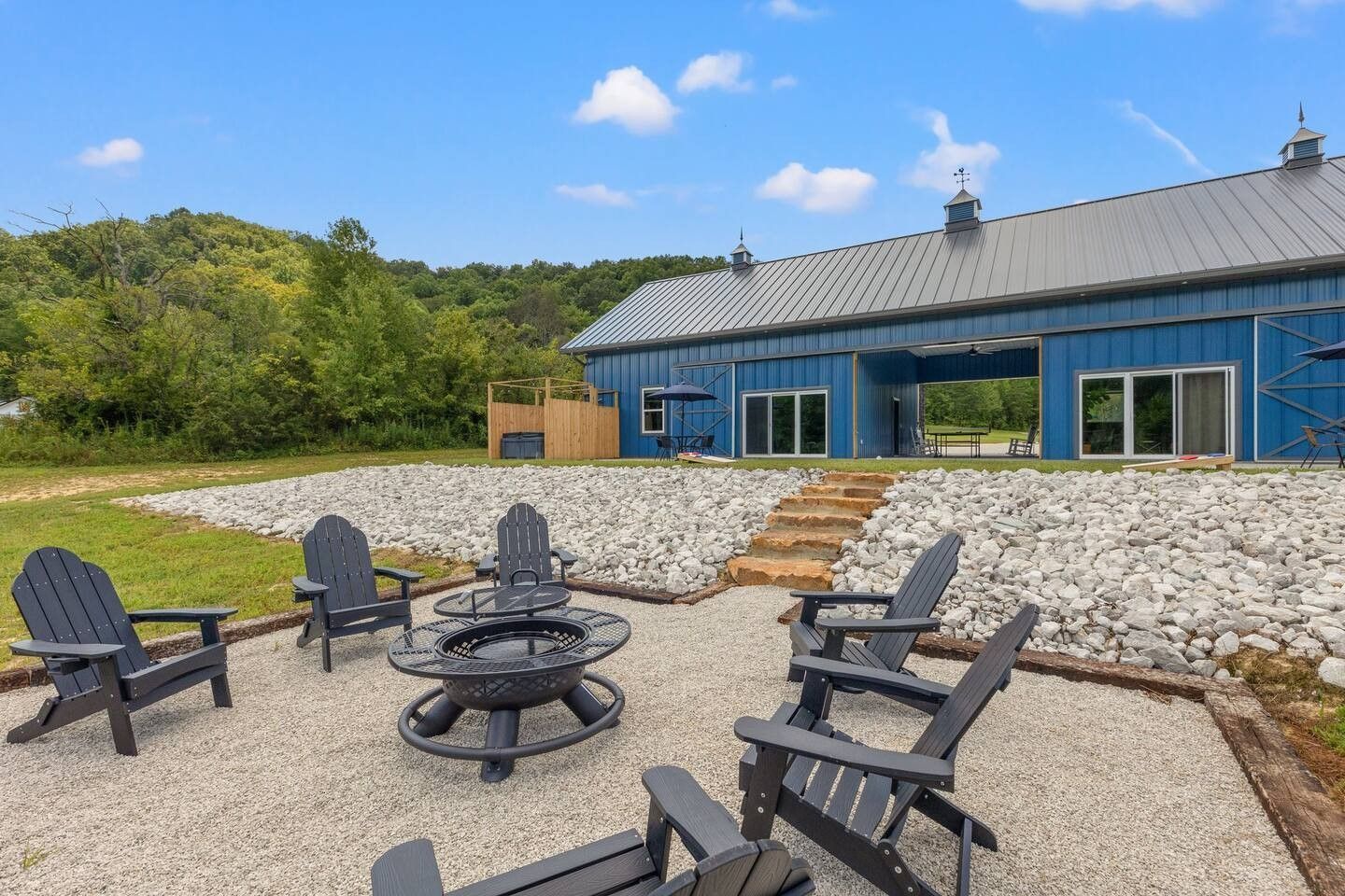 Blue barn-style house with a gravel patio, black Adirondack chairs, and a fire pit overlooking a grassy hillside.